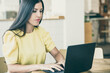 © Mangostar - Focused beautiful black haired woman sitting at table in co-working space, using laptop, looking at display and typing. Medium shot. Wireless communication or workplace concept