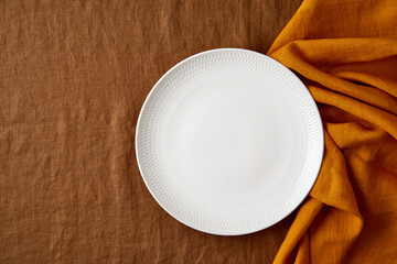 Clean empty white plate, fork and knife on bright dark brown tablecloth on table, yellow mustard napkin, copy space, mock up, top view. Concept for menu with utensil