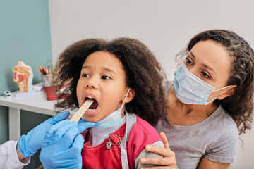 Wall Mural - Doctor using inspection spatula to examine African American girl's throat. Little girl with her mom at pediatrician appointment