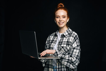 Smiling charming young business woman or student holding laptop computer and looking at camera on isolated black background. Pretty redhead lady model emotionally showing facial expressions