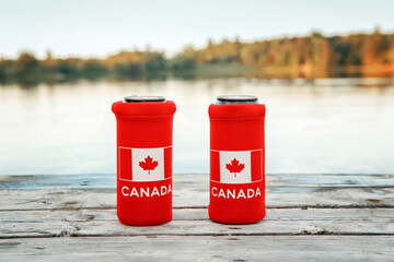 Wall Mural - Two cans of beer in red cozy beer can cooler with Canadian flag standing on wooden pier by lake outdoors. Friends celebrating Canada Day national celebration on July 1 in nature park by water.