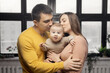 © Parilov - Happy family parents kiss small baby son on background of window, sunlight
