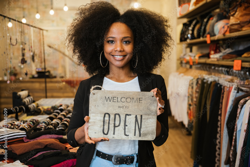 Portrait of the owner of the clothing store holding the sign with the words "Welcome we're open" in her hand to be attached to the window - Millennial woman welcomes customers at the entrance - obrazy, fototapety, plakaty 