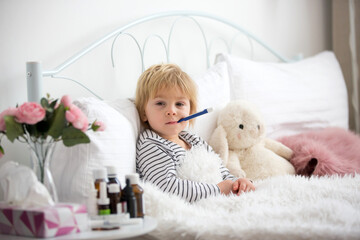 Sick little blond toddler boy, lying in bed with fever, hugging little fluffy toy, medicine next to him