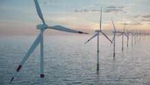 Camera Pulls Back To Show An Aerial View Of A Long Row Of Offshore Wind Turbines In The Sea Against Low Sun. Green And Renewable Energy Concept. Realistic High Quality 3d Animation. 