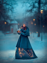 Girl in the blue cape standing at the night winter park with the snow ball