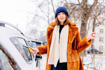 Winter portrait of a mature woman in red fur coat faux fur cleaning snow from a car. Beauty red haired senior female smiling s and cheerfully cleans the snow. Beautiful elderly woman outdoors.