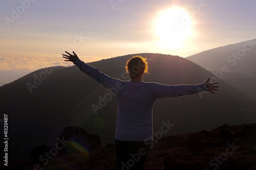 Rear View Of Woman With Arms Outstretched Standing On Mountain Against Sky During Sunset