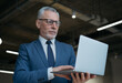 © ARUTA Images - Portrait of pensive grey haired businessman using laptop computer, typing, searching online, reading news at workplace. Handsome senior man wearing suit and glasses working in modern office