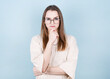 © MP Studio - Thoughtful model in the studio, in a beige T-shirt, glasses and a chain around her neck, looking to the side. hand near the face. isolated blue background
