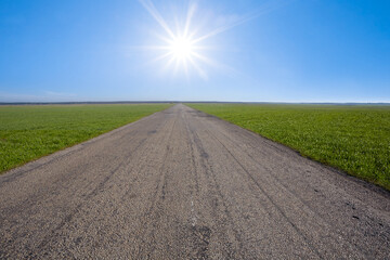 aspfalt road among green rural fields leaving far under a sparkle sun, transportation concept background