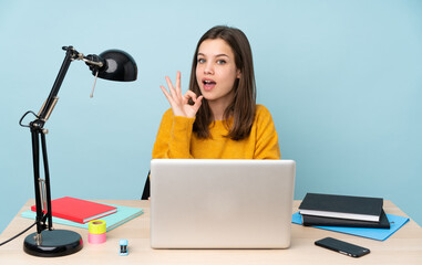 Student girl studying in her house isolated on blue background surprised and showing ok sign