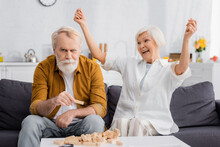 Excited Senior Woman Looking At Husband Holding Part Of Blocks Wood Game