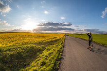 The Cyclist Stands On The Road And Looks Into The Distance And Admires The Beautiful Landscape At Sunset