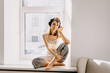 © Bostan Natalia - Young woman sitting by the window, listening to music in wireless headphones.
