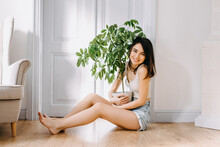 Young Woman Sitting On The Floor At Home, Hugging A Plant.