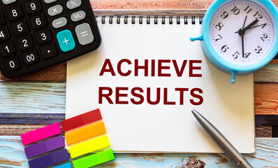 ACHIEVE RESULTS text written on a notebook, next to a calculator, pen, alarm clock and stickers.