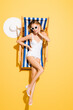 flirty woman in swimsuit and sunglasses touching face while posing in deck chair on yellow.