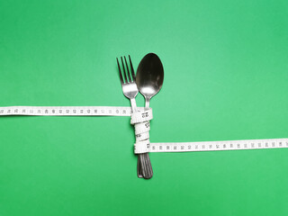 Spoon and knife are wrapped in white measuring tape on green background. Diet eating concept.Copy space for text.