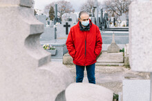Man Wearing A Mask, Sad And Praying, In Front Of The Grave Of A Deceased Relative In Times Of Covid-19