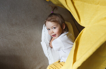 A beautiful girl in yellow pants and a white T-shirt smiles and plays inside the teepee at home on the couch. Top view.