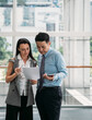 © Tijana - Two business people reading papers together in modern office with big windows.Two serious business people standing and talking about documents in office lobby