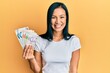 © Krakenimages.com - Beautiful hispanic woman holding singapore dollars banknotes looking positive and happy standing and smiling with a confident smile showing teeth