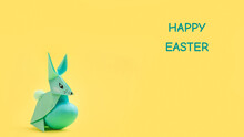 Paper Easter Bunny-origami Made Of Colored Paper And Painted Eggs On A Delicate Yellow Background. The Concept Of The Celebration Of Easter, Greeting Card, Crafts With Your Own Hands.