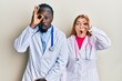 Young interracial couple wearing doctor uniform and stethoscope doing ok gesture shocked with surprised face, eye looking through fingers. unbelieving expression.