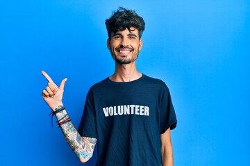 Young hispanic man wearing volunteer t shirt cheerful with a smile on face pointing with hand and finger up to the side with happy and natural expression