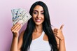 © Krakenimages.com - Beautiful hispanic woman holding dollars smiling happy and positive, thumb up doing excellent and approval sign