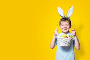 Wall Mural - Little cute boy with an Easter basket in his hands and bunny ears on his head on a yellow background. Easter background with place to insert text