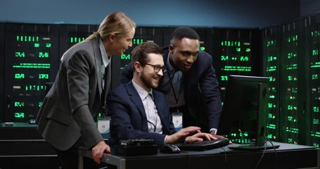 Two male programmers and one female system administrator stand over the workplace, look at computer and discuss tasks in server room.