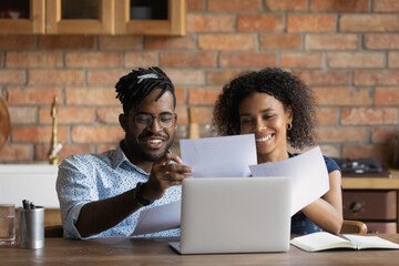 Close up happy African American family reading documents, holding papers, using laptop, wife and husband calculating domestic bills, planning budget, browsing online banking service in kitchen