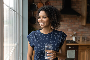 Wall Mural - Close up dreamy smiling African American woman holding glass of pure fresh mineral water, standing near window in kitchen, starting new day, healthy lifestyle habit, body and skin care concept