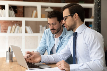 Canvas Print - Focused young Caucasian male colleagues look at laptop screen brainstorm cooperate discuss business project together. Serious men coworkers employees work on computer. Collaboration concept.