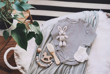 Still life background of cute baby products - changing basket with baby bodysuit, newborn clothes, knitted rabbit and wooden toy. Minimalist style photography of baby shower, pregnancy announcement.