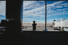 Rear View Of Silhouette Baby Boy Looking Through Window At Airport Departure Area