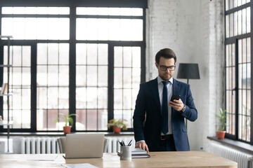 Poster - Serious young Caucasian businessman in suit stand near office desk look at cellphone screen consult client online. Focused male CEO or boss use modern smartphone gadget for business negotiation.