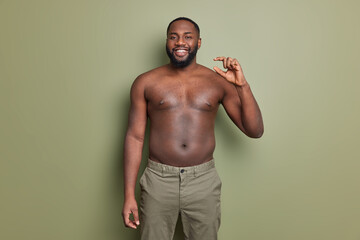 Wall Mural - Glad dark skinned man poses with bare torso shapes little gesture tells everyone about his poor salary measures small invisible object. Happy African American guy demontrates tiny size of something