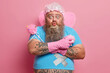 © wayhome.studio  - Funny bearded man keeps arms crossed wears rubber gloves holds sponge and magic wand pouts lips as wants to kiss you ready for cleaning poses with household supplies isolated over pink background