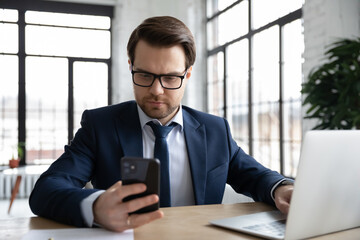 Canvas Print - Concentrated Caucasian businessman work on laptop and cellphone online in office. Serious male CEO or employee busy browsing internet on computer, consult business client on smartphone on web.