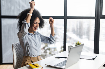 african-american woman with curly hairstyle hands up,at the desk with open laptop,wide smile,excited