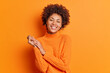 © WHstudio Leushin N - Pretty curly haired young woman smiles gently keeps hands together focused directly at camera has pleasant talk with best friend dressed in casual sweater isolated over vivid orange background