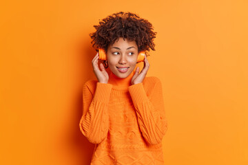Wall Mural - Thoughtful pretty millennial female with Afro hair listens music concentrated aside wears wireless headphones on ears dressed in long sleeved casual sweater isolated over orange studio background