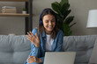 © fizkes - Smiling teenage female pupil sit on sofa in headphones wave hand to laptop screen greet teacher tutor at lesson consultation. Happy young woman say hi hello to friend speaking from home by video call