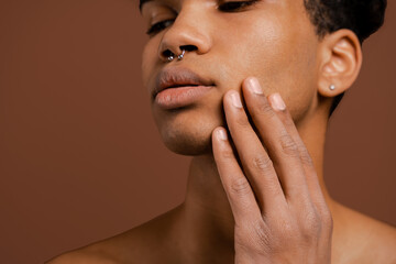 Photo of attractive black man with piercing touches his smooth face. Naked torso, isolated brown color background