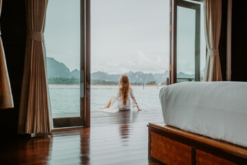 Travel Woman in White Dress Sitting Outdoor near Wooden House on Water. View on Tourist Woman Relaxing and Leisure from Raft House on Cheow Lan Lake in Thailand. Eco Tourism