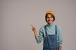 © Ivan - Photo of cute female hipster smiles and points at left side. White woman wears denim overall and hat isolated grey color background