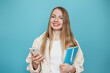 © Evgenia - Blonde student girl holding mobile phone and notepad looking at the camera and smiling isolated on blue studio background. Online education